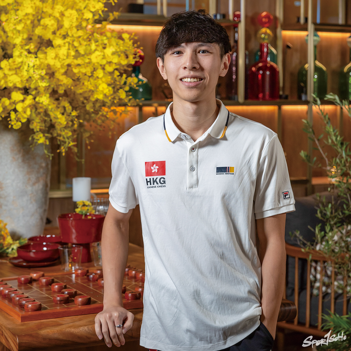 Tony Fung in a white Hong Kong team shirt, one hand resting on the edge of a xiangqi board, smiling at the camera.