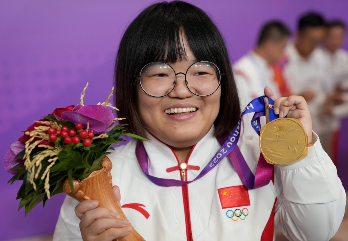 Zuo Wenjing in a white China team jacket, smiling through round glasses, holding flowers in one hand and her gold medal up in the other.